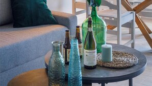 Un salón acogedor con un sofá, una mesa redonda de centro y una mesa de comedor con sillas. Hay elementos decorativos como un jarrón con flores, una botella de vino y una vela sobre la mesa de centro. Se ve también una lámpara con pantalla roja y una lámpara colgante.