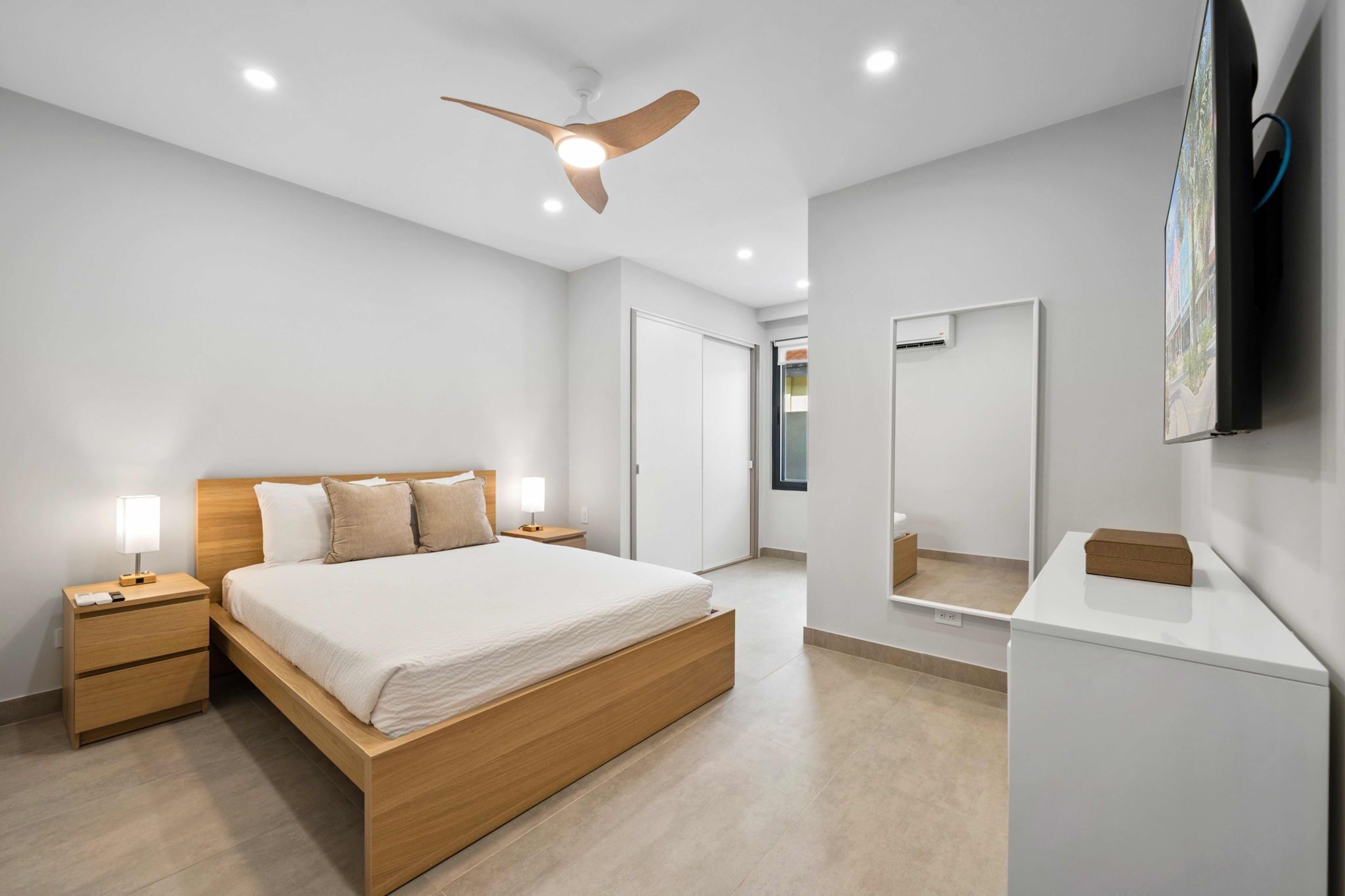 A modern bedroom with a wooden bed, bedside tables with lamps, a ceiling fan, a large mirror, and a TV mounted on the wall.