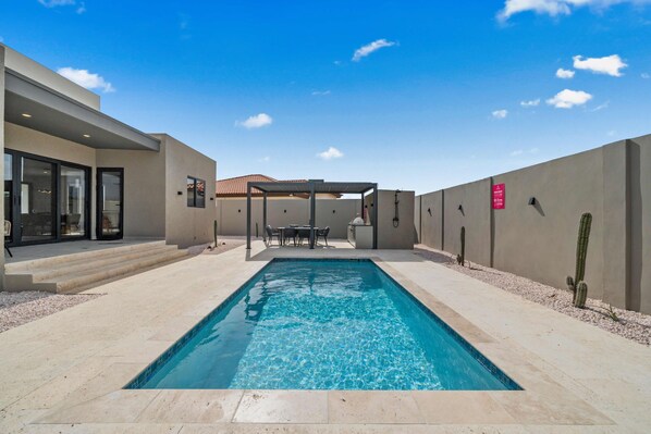 A rectangular swimming pool with clear blue water, surrounded by a patio area with steps leading to a covered seating area. - Safir 40 (Noord)