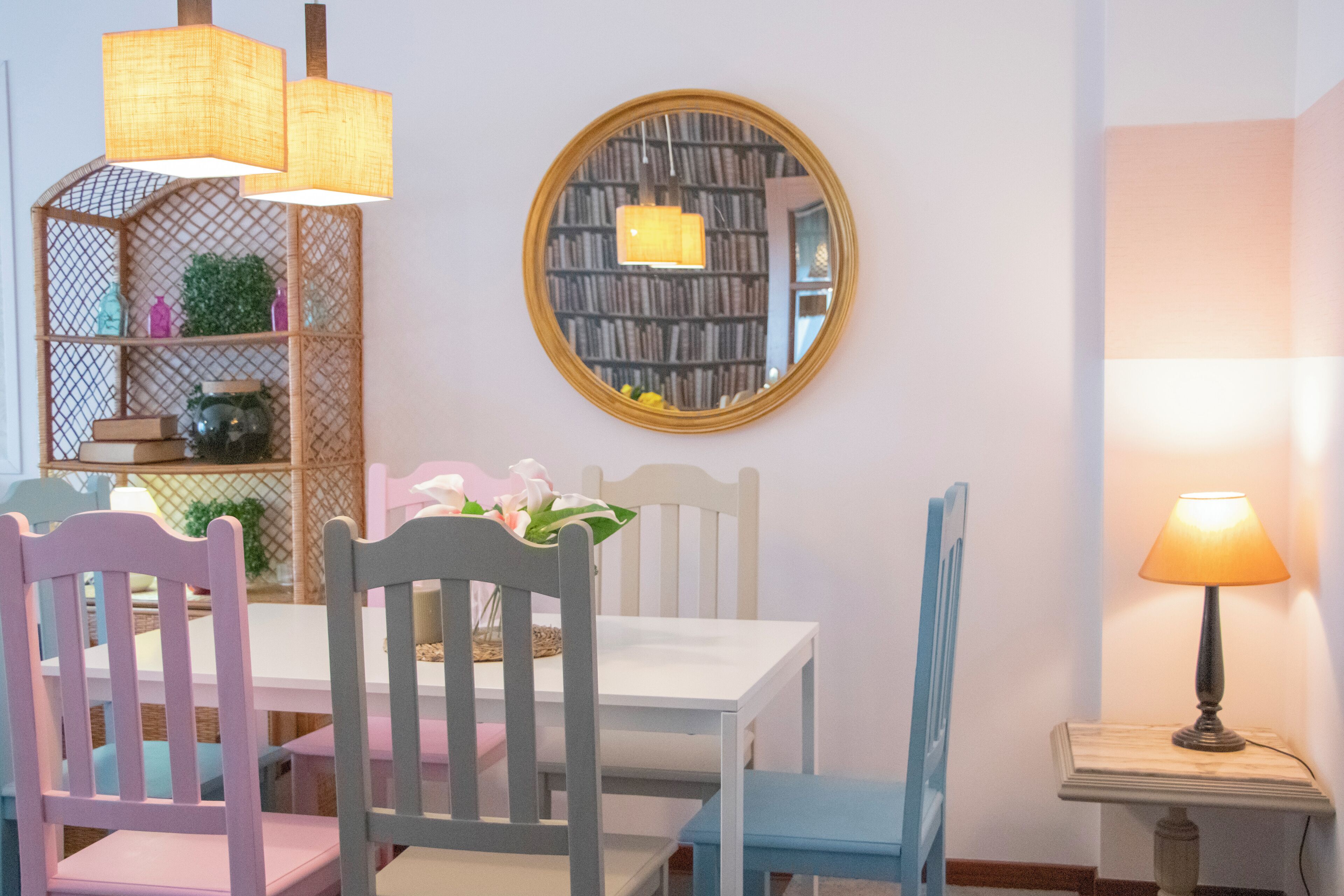 Un coin repas avec une table blanche, des chaises bleues et roses, un miroir rond et une bibliothèque contenant des livres et des objets décoratifs.