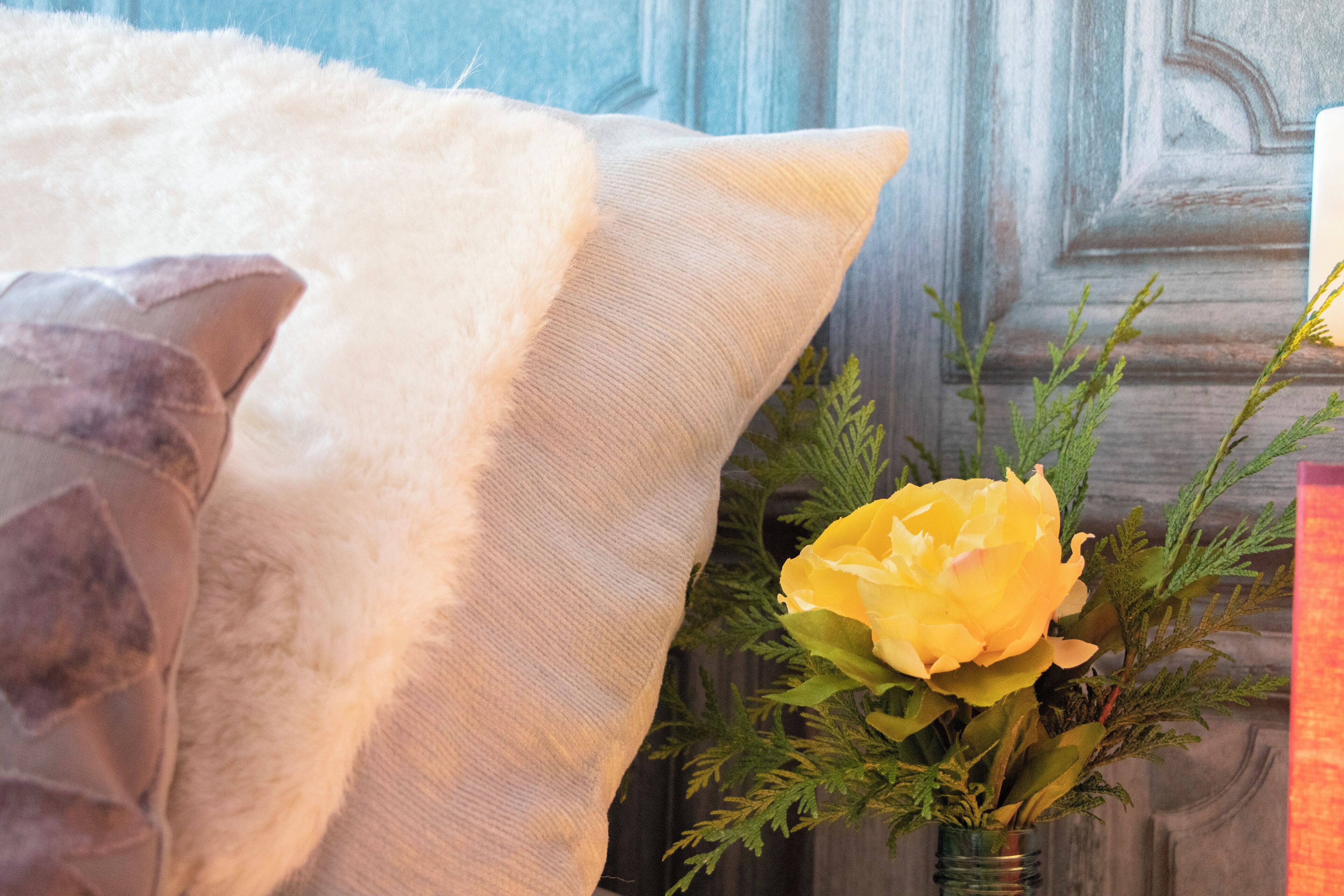 Un lit avec des oreillers blancs et gris, une fleur jaune dans un vase en verre et, au fond, une porte en bois.