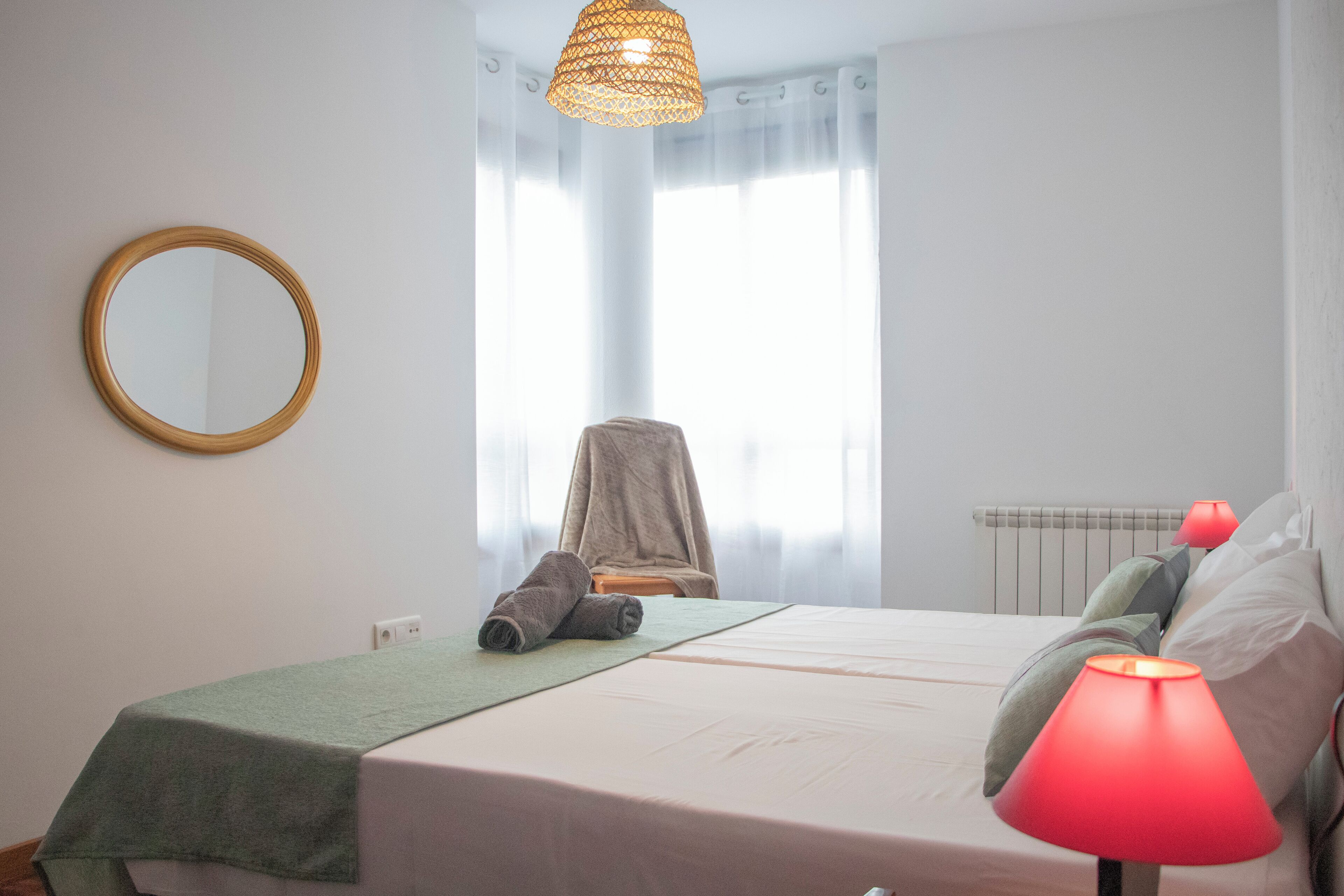 A bedroom with a bed, a round mirror, a chair, a red lamp, and a window with curtains.