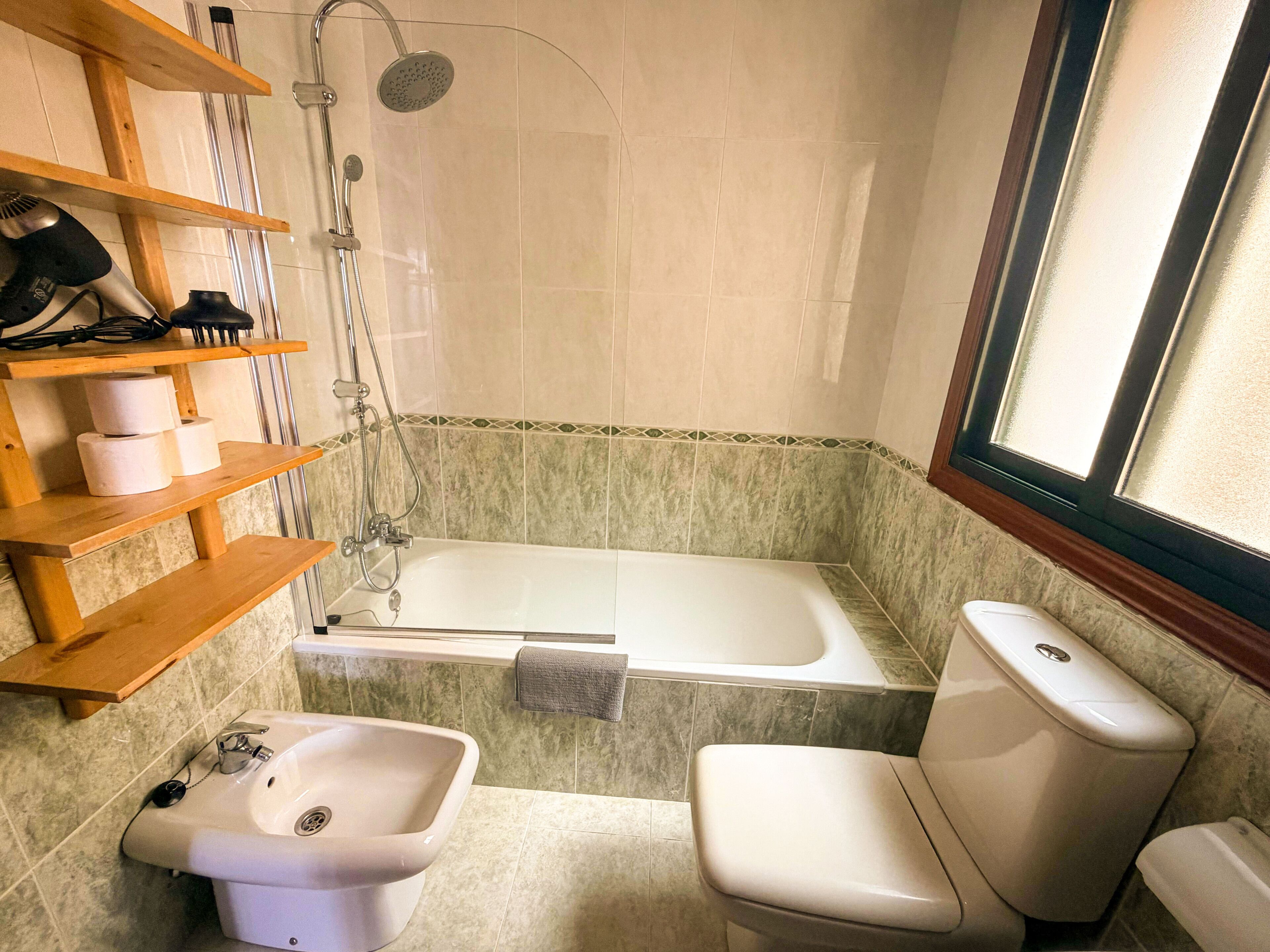 Baño con bañera, lavamanos, inodoro y ducha. Hay repisas con artículos de aseo y una ventana que proporciona luz natural.