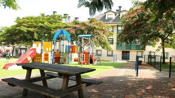 Ein Spielplatz mit verschiedenen Spielgeräten, einem Picknicktisch und einem Gebäude im Hintergrund.