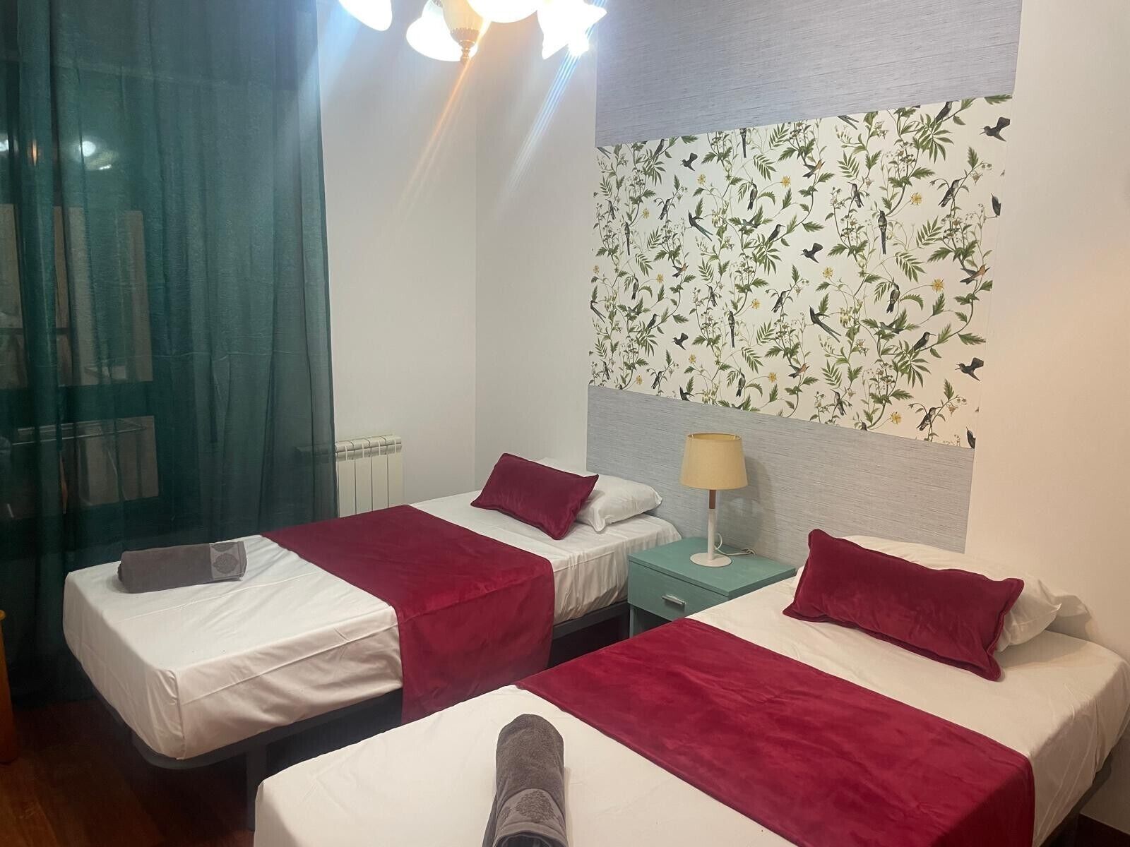 A hotel room with two beds, a window with curtains, a radiator, and a wall with a floral wallpaper.