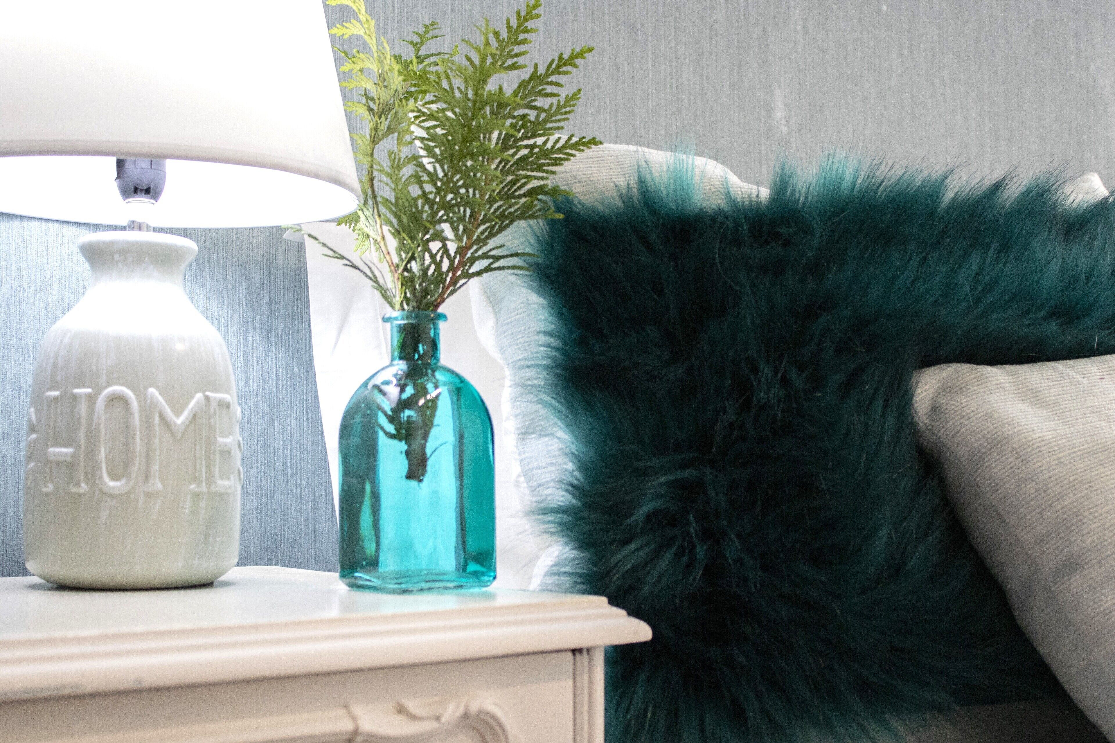 A white bedside table with a white lamp and a glass vase with green foliage.