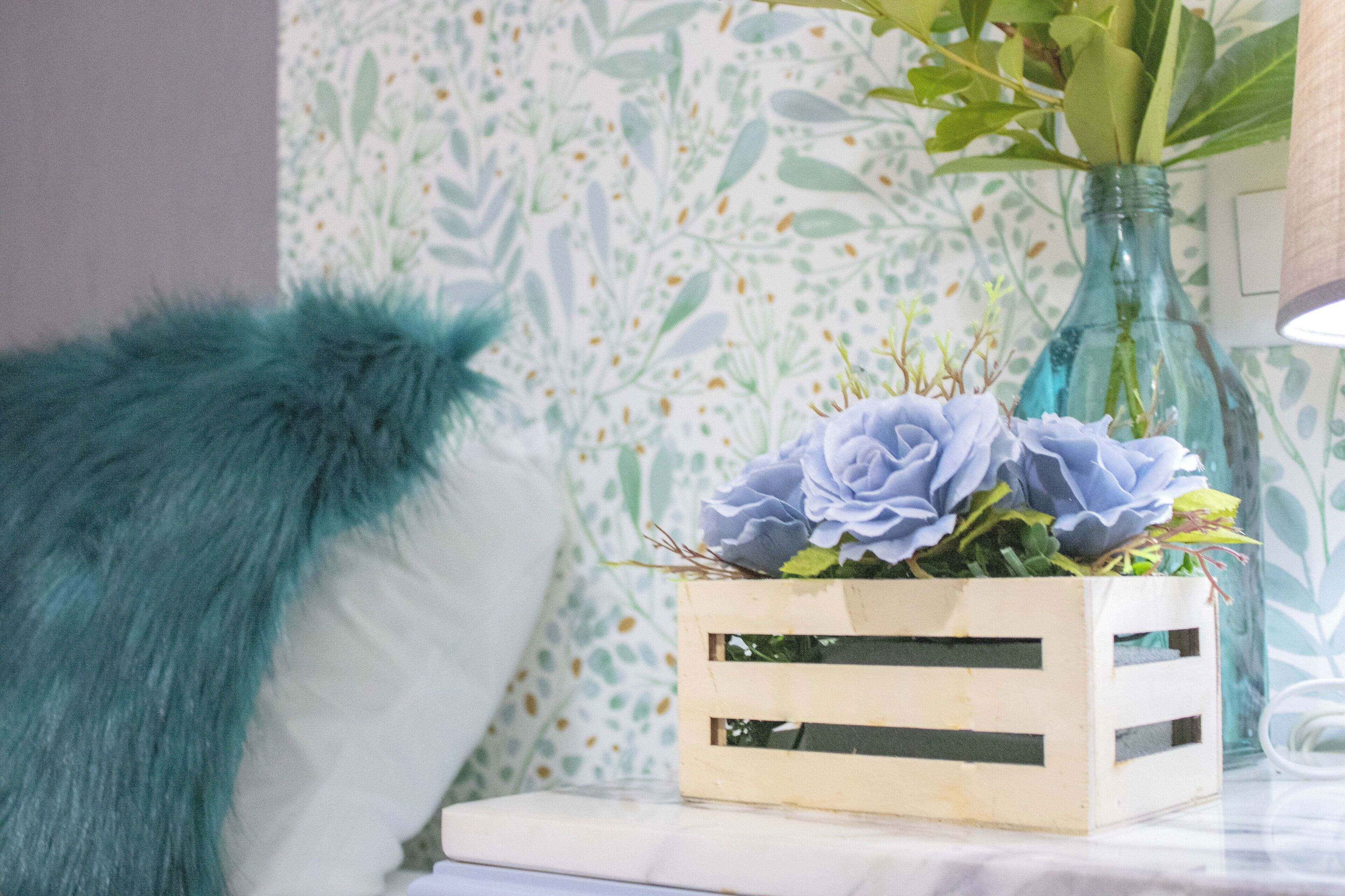 Ein Holzkoffer mit einer Blumenkomposition auf weißer Oberfläche, ein flauschiges blaues Kissen und ein Hintergrund mit gemustertem Tapetenmuster.