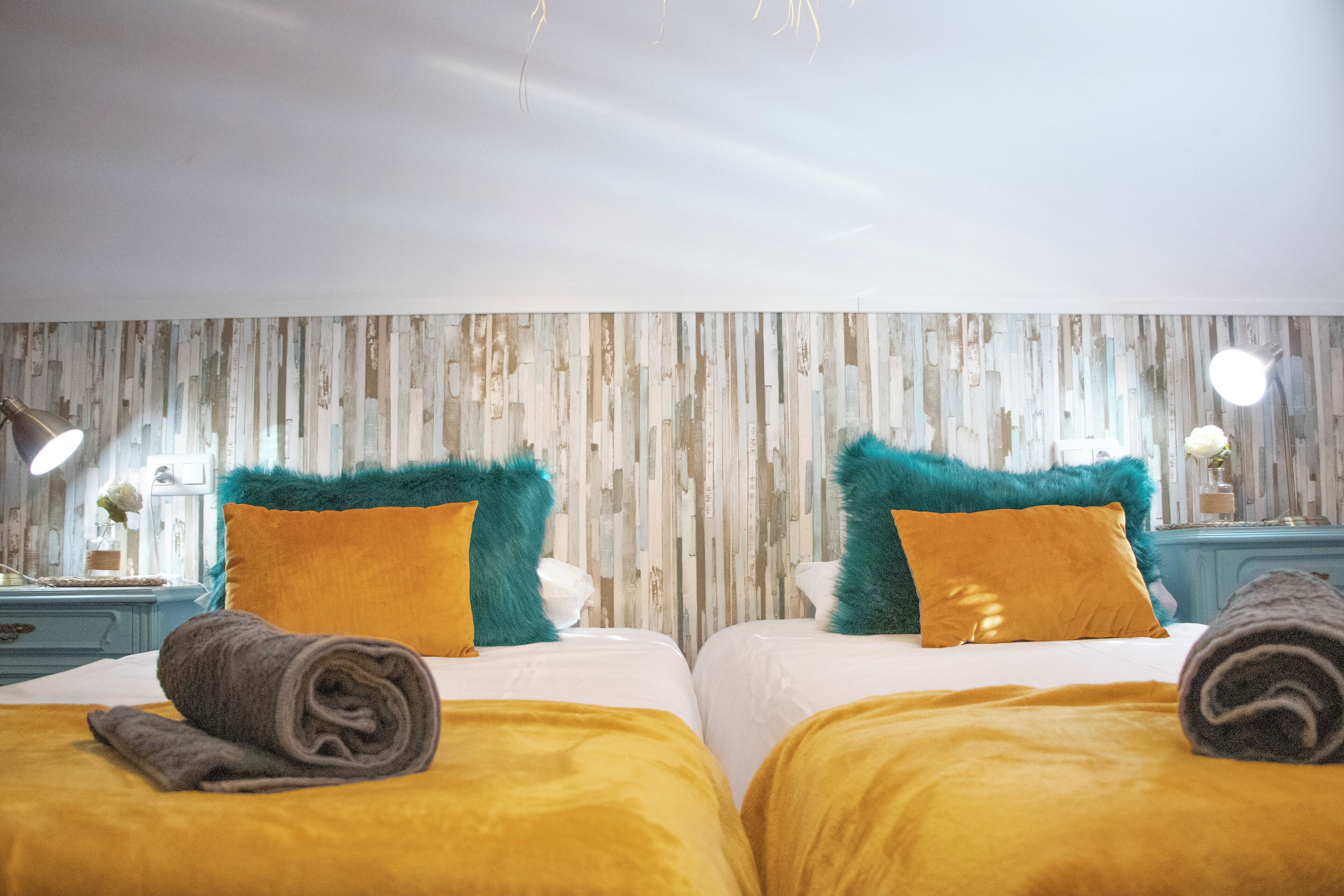 Habitación de hotel con dos camas, cada una con cubrecama amarillo y una toalla enrollada. La habitación tiene paneles de madera y una mesita de noche con una lámpara y un florero.
