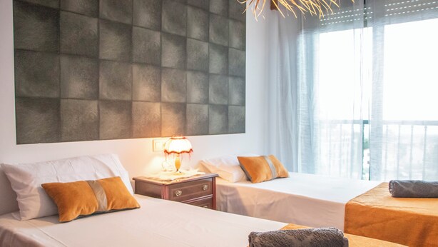 Une chambre d’hôtel avec deux lits, une grande tête de lit rembourrée, une table de chevet avec une lampe et une vue sur l’extérieur à travers des rideaux légers.