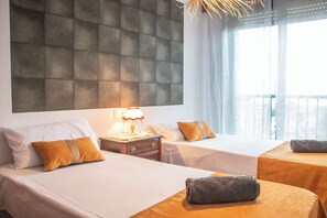 Ein Hotelzimmer mit zwei Betten, einem großen, gepolsterten Kopfteil, einem Nachttisch mit Lampe und Blick ins Freie durch durchsichtige Vorhänge.