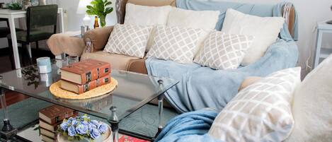 A cozy living room with a sofa, a glass coffee table, and a side table with books and a lamp.