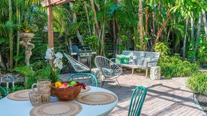 An outdoor patio with a round table, chairs, a grill, and lush tropical plants.