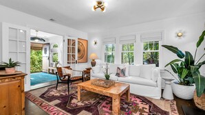 A spacious living room with a white sofa, a wooden coffee table, and a patterned rug. There are large windows with views of greenery outside.