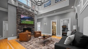 A spacious living room with a stone fireplace, a black leather sofa, and a large TV mounted on a stone wall.