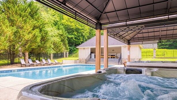 A hot tub area with a pool and lounge chairs, surrounded by trees and a covered patio.