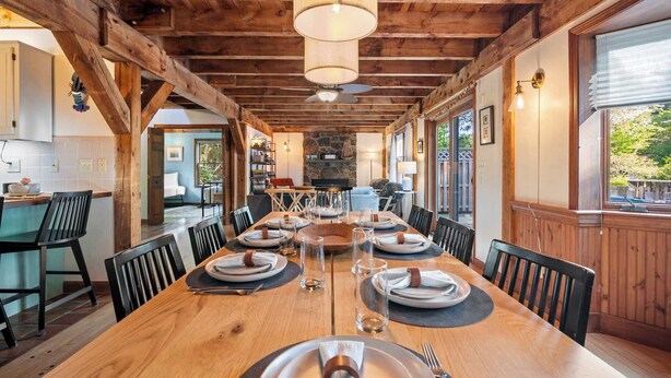 Une salle à manger en bois avec une longue table dressée pour six personnes, agrémentée d’un lustre, d’un mur en pierre et de grandes fenêtres.
