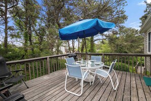 Een houten terras met een ronde glazen tafel, vier stoelen, een blauwe parasol en een barbecue.