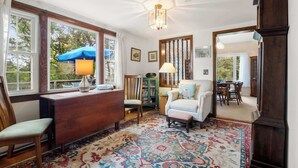 A cozy room with a wooden desk, a chair, a patterned rug, and a view of the outdoors through large windows. - Cozy Cottage Walk to Nauset Beach! (Orleans)