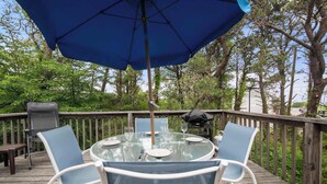 A round glass table with four chairs, a blue umbrella, and a wooden deck with railings. - Cozy Cottage Walk to Nauset Beach! (Orleans)