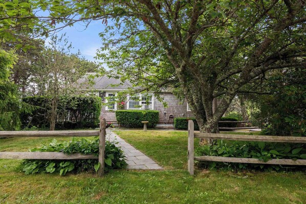 Un chemin de jardin menant à une maison à la façade en pierre, entourée d’une végétation luxuriante et d’une clôture en bois.