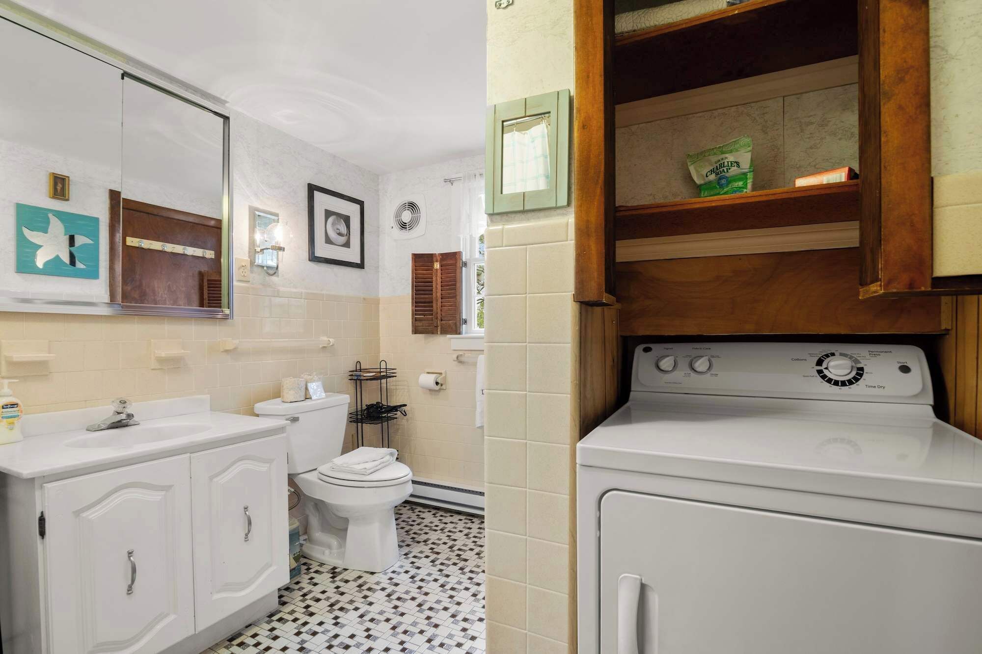 A bathroom with a white sink, toilet, and a washing machine. There are tiled floors and a mirror above the sink.