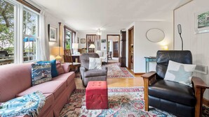 A spacious living room with a large window, a fireplace, and a cozy seating area with a red ottoman. - Cozy Cottage Walk to Nauset Beach! (Orleans)