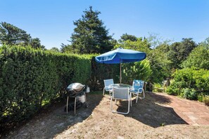 A patio with a table and chairs under a blue umbrella, surrounded by dense hedges and trees. - Studio, Walk to Nauset Beach! (Orleans)