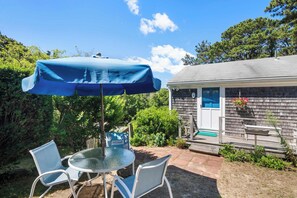 A patio with a round glass table and two chairs, a blue umbrella, and a house with a blue door and a hanging flower basket.