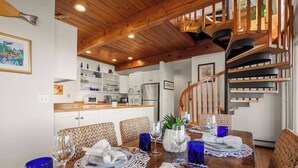 A dining area with a wooden table set for two, a kitchen in the background, and a spiral staircase.