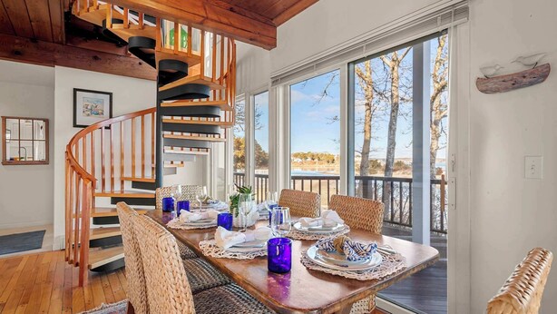A dining area with a wooden table set for four, surrounded by wicker chairs, a spiral staircase, and large windows offering a view of trees and a body of water.