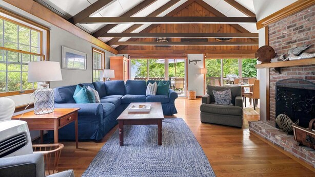 A spacious living room with a large blue sectional sofa, a fireplace, and a view of the outdoors through large windows. - Great location with Water Views! (Harwich)