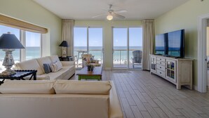 A spacious living room with a large television, a sofa, a coffee table, and a view of the ocean through large windows. - 401E Beach Service Included! End Unit! (Panama City Beach)