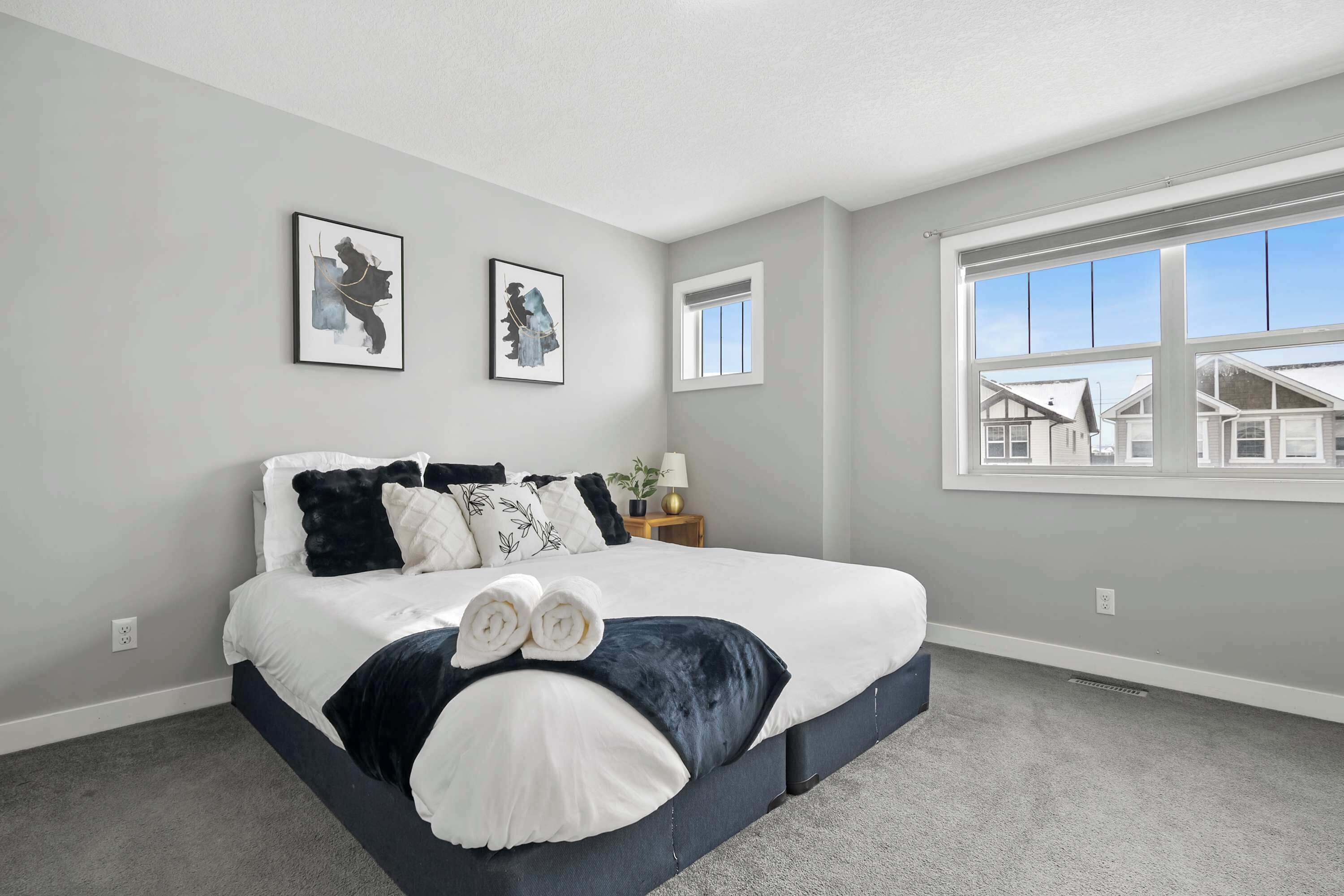 A bedroom with a large bed, two framed artworks on the wall, a window with a view of houses, and a small bedside table with a lamp.
