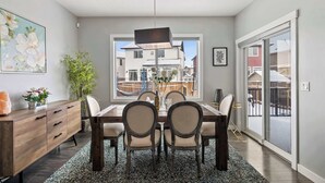 A dining area with a wooden table, four chairs, a dark pendant light, a large window with a view of neighboring houses, and a sliding glass door.