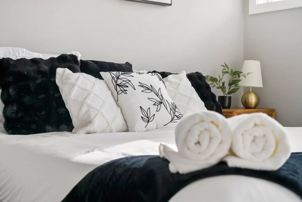 Una cama bien hecha con toallas enrolladas, almohadas blancas y negras y una planta en la mesita de noche.