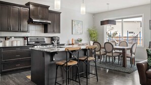 A modern kitchen with dark cabinetry, a central island with bar stools, a dining area with a table and chairs, and a view of the outside through a large window.