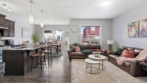 A modern kitchen with dark wood cabinets, a central island with bar stools, and pendant lighting. A living area with a brown leather couch, a round coffee table, and a large rug is visible.