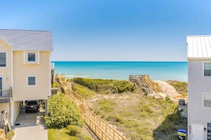 A beachfront property with a wooden staircase leading to a sandy area, a clear view of the ocean, and a building with a white facade.