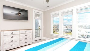 A room with a flat-screen TV mounted on the wall, a ceiling fan, a wooden dresser with drawers, and a view of a residential area through large windows. - Dune Just Fine - Gorgeous 5BR Home - Pool, Elevator, Porches & More! (Surf City)
