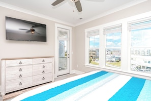 A room with a flat-screen TV mounted on the wall, a ceiling fan, a wooden dresser with drawers, and a view of a residential area through large windows.