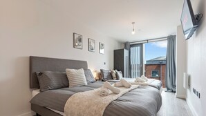 Una camera da letto moderna con un letto grande, biancheria grigia e una soffice coperta bianca. Si notano tre quadri appesi al muro e un televisore montato a parete.