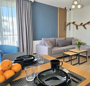 Una sala de estar moderna con un sofá gris, una mesa de centro de madera y una mesa de comedor dispuesta con platos, cubiertos y un florero con flores amarillas.