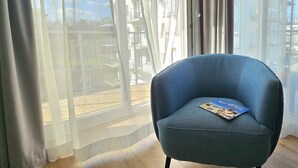 A blue armchair with a magazine, positioned near a window with sheer curtains, overlooking a building with balconies.