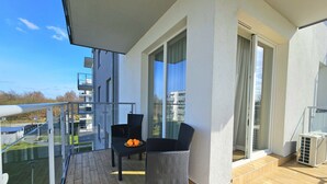 A balcony with two wicker chairs, a small table with oranges, and a glass railing overlooking a residential area.