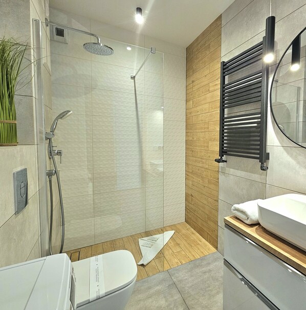 Un baño moderno con ducha de vidrio, inodoro blanco, tocador de madera con espejo y lavamanos blanco.