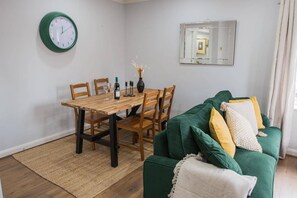 A dining area with a wooden table, chairs, and a green sofa with cushions. A wall clock and a mirror are also visible. - Sienna Haven Sleeps 4 With Parking (Birmingham)
