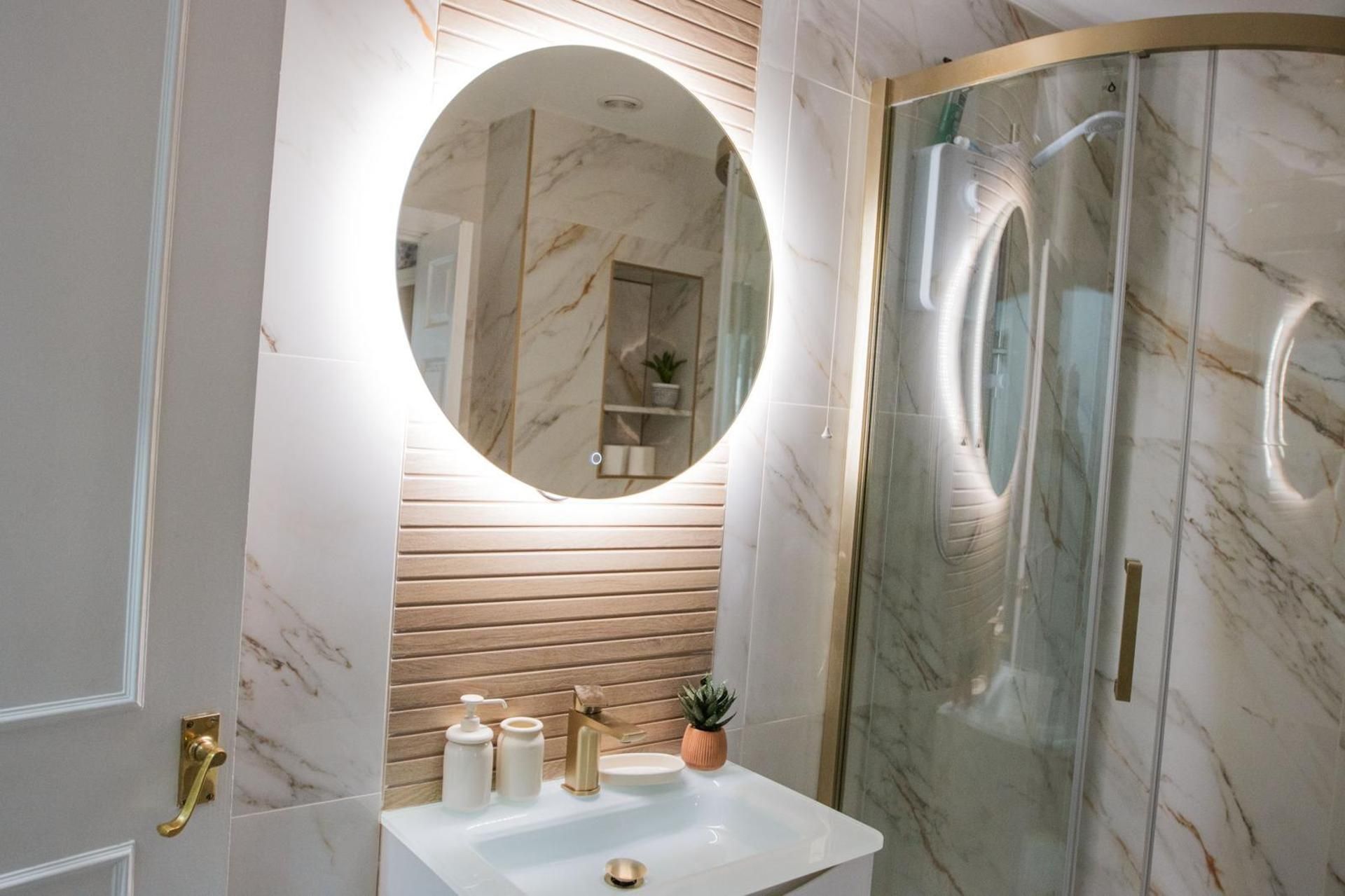 A bathroom with a marble sink, a round mirror, a glass shower enclosure, and wooden paneling.