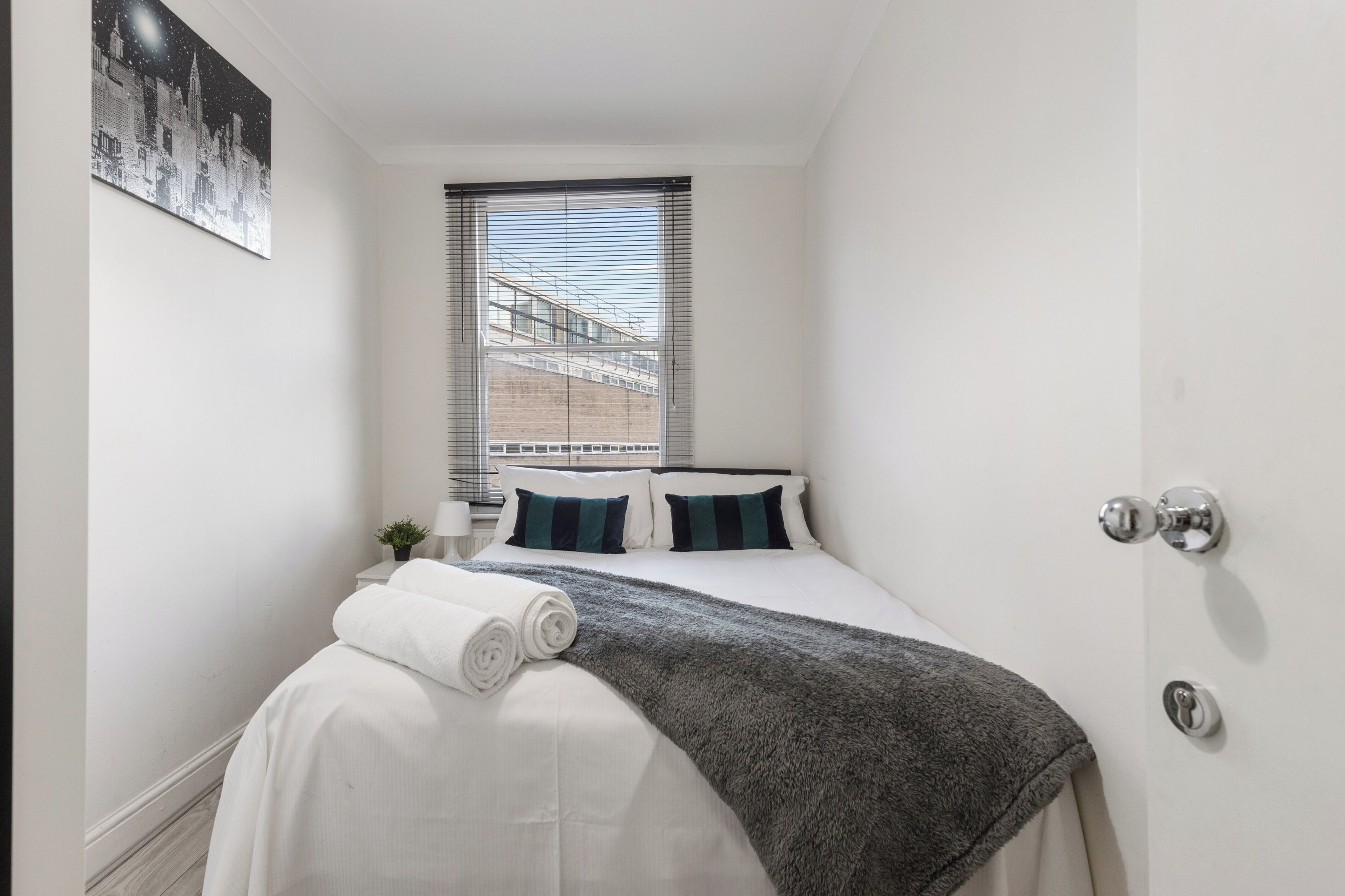 Una cama bien hecha con sábanas blancas y un plaid gris, una ventana con persianas, y un cuadro de un paisaje urbano en la pared.