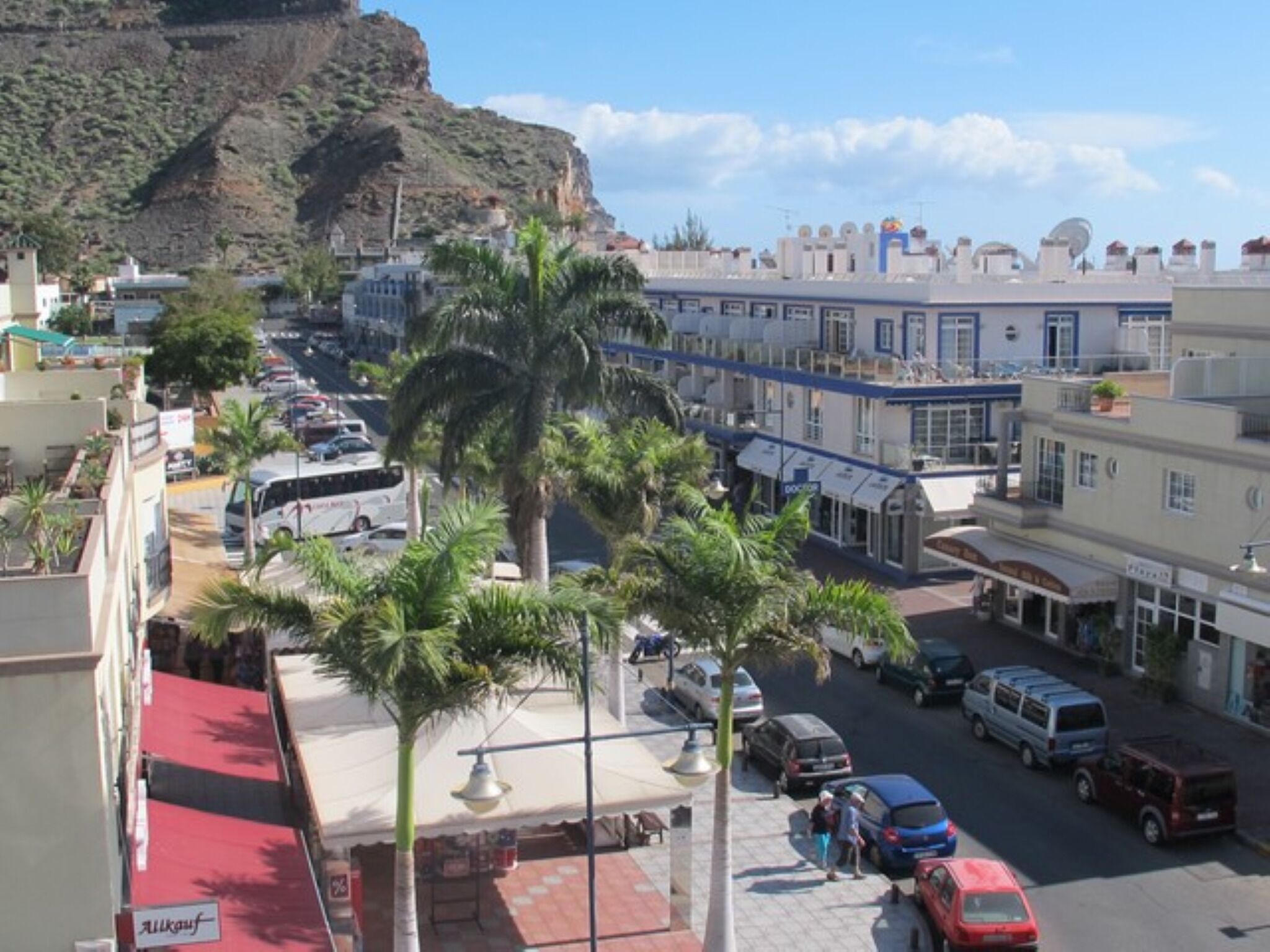 A street view with buildings, parked cars, and palm trees.