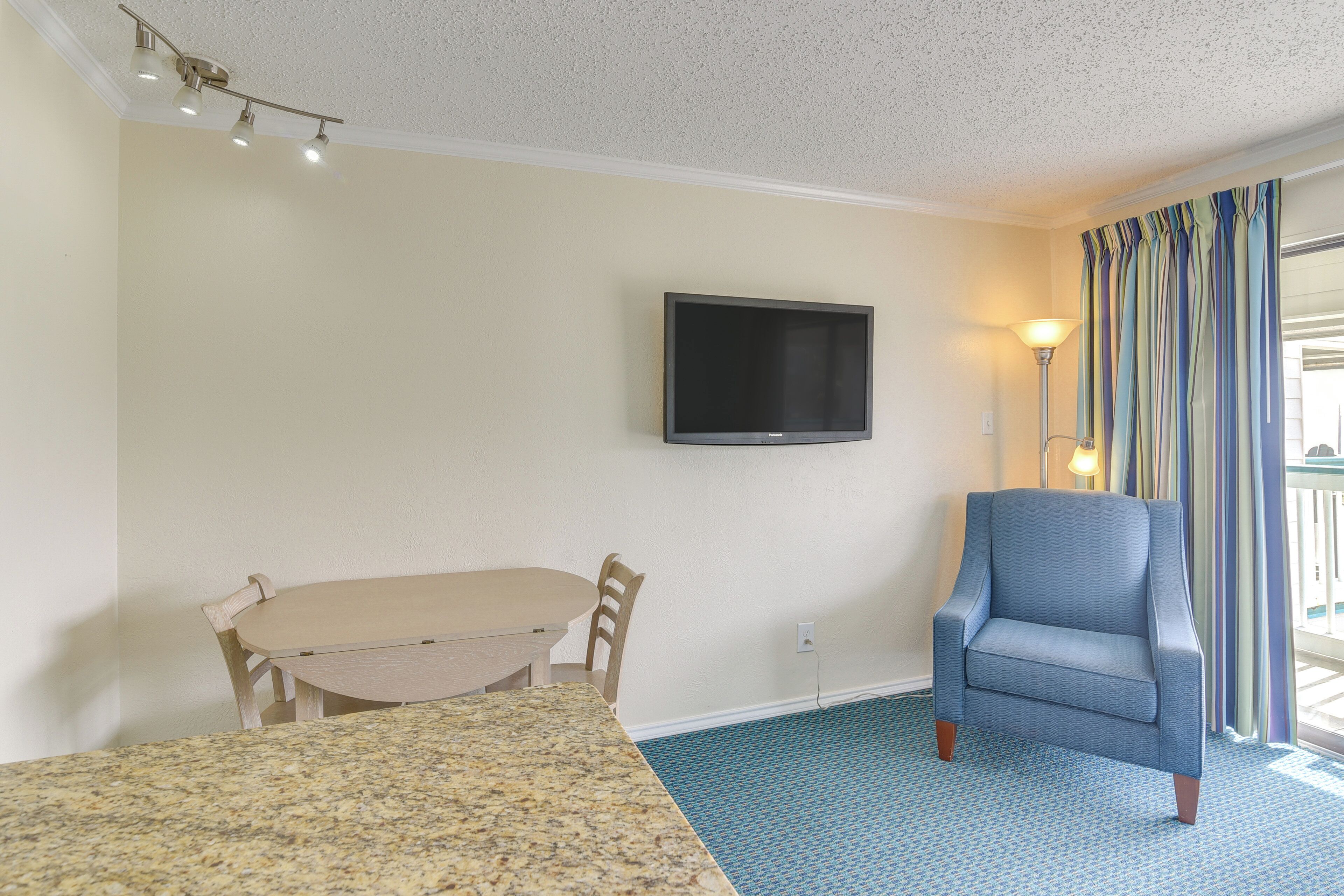 A hotel room with a television mounted on the wall, a dining table with chairs, a blue armchair, and a patterned carpet.